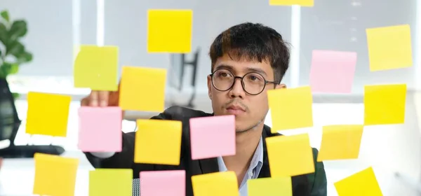 Young asian businessman writing on sticky note at office, Business brainstorming creative ideas, Asian office man writing adhesive note on glass wall, Business strategy planning to success concept