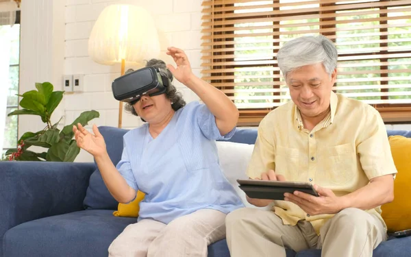 Senior asian couple playing virtual reality headset and using digital tablet in home living room with happiness, Asia elderly retirement old people lifestyle and technology