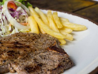 T-bone Steak tuz ve biber, altın kızarmış patates ve yeşil sebzeler ile kapatın. Beyaz tabak, çatal ve bıçak, restoranda ahşap masa