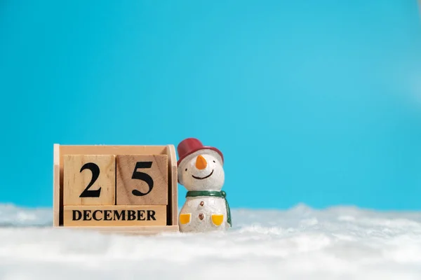 Snowman wearing red hat sitting beside wooden block calendar set on the Christmas date 25 december on white wool and blue background. Copy space for text or content. Concept of merry christmas.