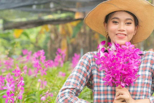 A woman gardener wearing a plaid shirt wearing a hat is holding a pink orchid in his hand. And was smiling happily. In the orchid garden