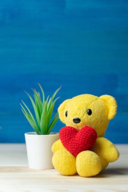 El yapımı kırmızı kalp beyaz tencere önündeki sarı oyuncak ayı ve ahşap masa ve mavi arka plandaki yeşil süslemeli bitkiler. Sevgililer Günü kavramı.