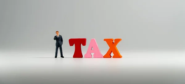miniature figure businessman in dark blue suit standing beside colorful wooden TAX word and thinking abount TAX.