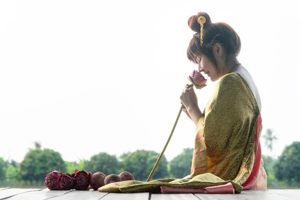 Schöne asiatische Frauen in traditioneller thailändischer Kleidung und auf Holzboden sitzend. ihre Hände halten Lotusblume und riechen rosa Blume. — Stockfoto