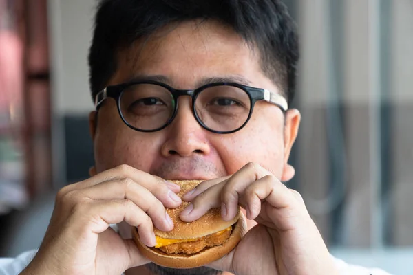 Asia man is eating hamburger in a fast food restaurant and enjoying delicious food. man in a White t-shirt and glasses holding a burger and enjoying the taste of hamburger