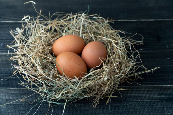 Close up of fresh brown chicken eggs in hay nest on black wooden background. Concept of organic eggs, free space for text or other elements