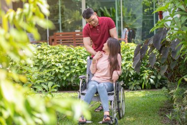 Asyalı kadın tekerlekli sandalyede ve kocasıyla gülümsüyor. Tekerlekli sandalyenin arkasında duran bir adam, bir kaza sonucu ayakları bacağını inciyen karısını cesaretlendiriyor. Bakım ve destek kavramı