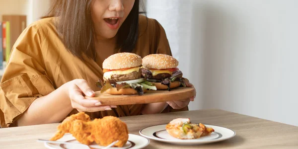 Aç kilolu kadın ahşap plaka üzerinde hamburger tutan, Kızarmış tavuk ve Pizza masada . Alem yeme bozukluğu kavramı (Yatak).