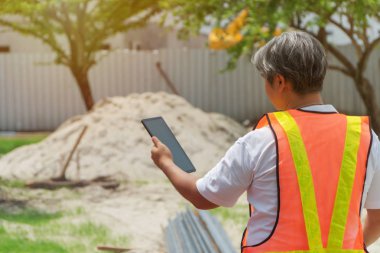 İnşaat alanının önünde beyaz bir kaskla duran mühendislik, inşaat için tablet kullanıyor. İnşaatın doğruluğunu kontrol etme ve kontrol etme kavramları