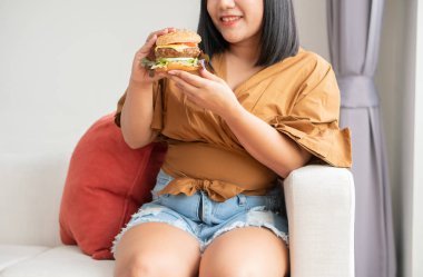 Hungry overweight woman smiling and holding hamburger and sitting in the living room, her very happy and enjoy to eat fast food. Concept of binge eating disorder (BED). clipart