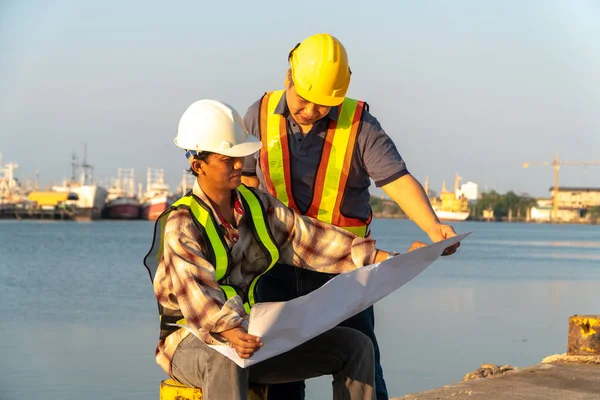 Two engineers stood wearing a safety helmet. Standing at the pier And holding the blueprint And consult the plan In the construction. Concept of efficient construction management.