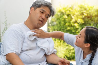 Orta yaşlı ve obez erkekler felçli, ağzı yamuk, elleri buruşuk ve elleri uzuvları kırık tekerlekli sandalyede ve kocasının yüzüne havluyu sevgiyle ve umutla kullanan bir kadın var.