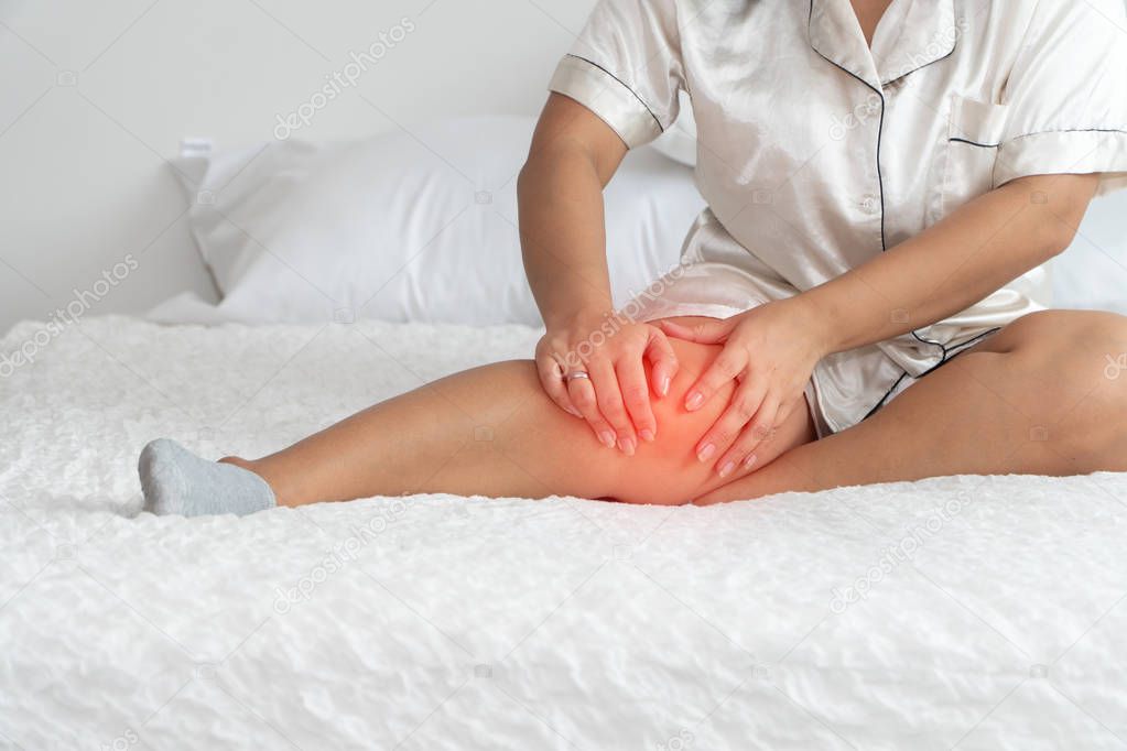 Overweight woman sitting on the bed and catching the knees because of the pain caused by overweight problems. Concept of health problems of obese people.