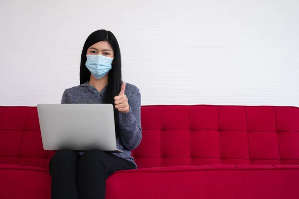 フェイスマスクを着用し ソファの上にラップトップを使用しているアジアの女性は 自宅やThumbsから働いています 隔離中の新しい生活習慣 家にいるという概念 — ストック写真