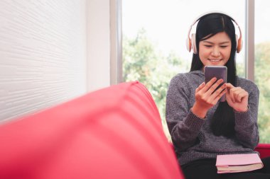 Tatlı Asyalı kadın sabahları kanepede oturuyor ve müzik dinlerken cep telefonlarına mesajlar gönderiyor ve kucağında bir kitapla oturma odasında.