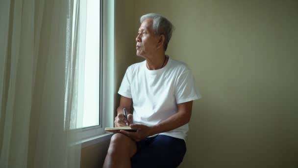 Šťastný starší muž sedí u okna a píše deník pro pokladníka zkušenosti z minulého života. — Stock video