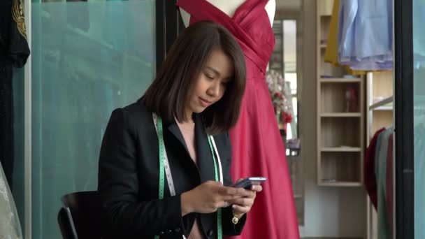 Feliz joven mujer asiática modista diseñador de moda utiliza un teléfono inteligente para aceptar pedidos del cliente en una sala de exposición. Concepto de joven empresario exitoso en el negocio de la moda . — Vídeos de Stock