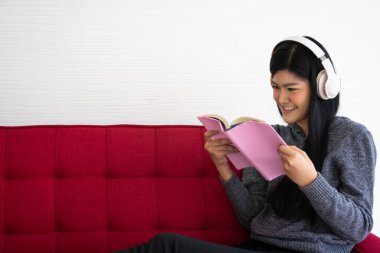 Tatlı Asyalı kadın sabahları kanepede oturuyor ve oturma odasında müzik dinlerken kitap okuyor.