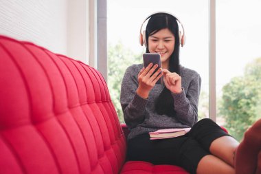 Tatlı Asyalı kadın sabahları kanepede oturuyor ve müzik dinlerken cep telefonlarına mesajlar gönderiyor ve kucağında bir kitapla oturma odasında.