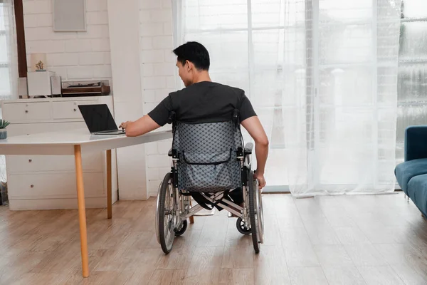 Happy disabled Asian man sitting in a wheelchair and working with a computer at home.
