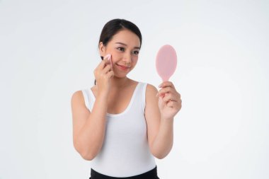 Güzel, genç Asyalı bir kadın pamukla cildini temizliyor ve elinde beyaz arka planda izole edilmiş makyaj aynası tutuyor.