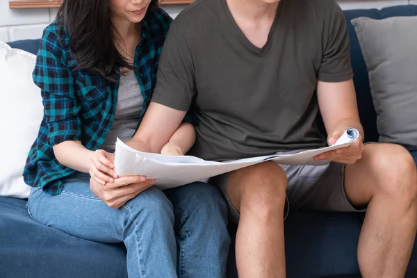 Happy young Asian couple sitting on the sofa and holding House plan for planning decoration in moving to a new house on the first day. Concept of starting a new life for a newly married couple.