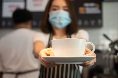 Bulaşıcı hastalıkları önlemek için yüz maskesi takan Asyalı garson kadın kahve fincanı tutuyor. COVID 19 salgınından korunma ve güvenlik kavramı