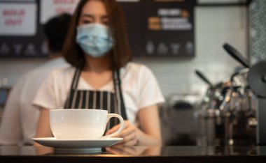 Kahve fincanının seçici odağı ve bulaşıcı hastalıkları önlemek ve kahve dükkanındaki müşterilere hizmet etmek için yüz maskesi takan Asyalı baristanın arka planı. COVID 19 'dan önleme kavramı