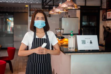 Asyalı garson kadın yüz maskesi takıyor ve elinde kızılötesi alın termometresi var. Restorana girmeden önce vücut ısısını ölçmek için müşterilerin semptomlarını kontrol ediyor. ).