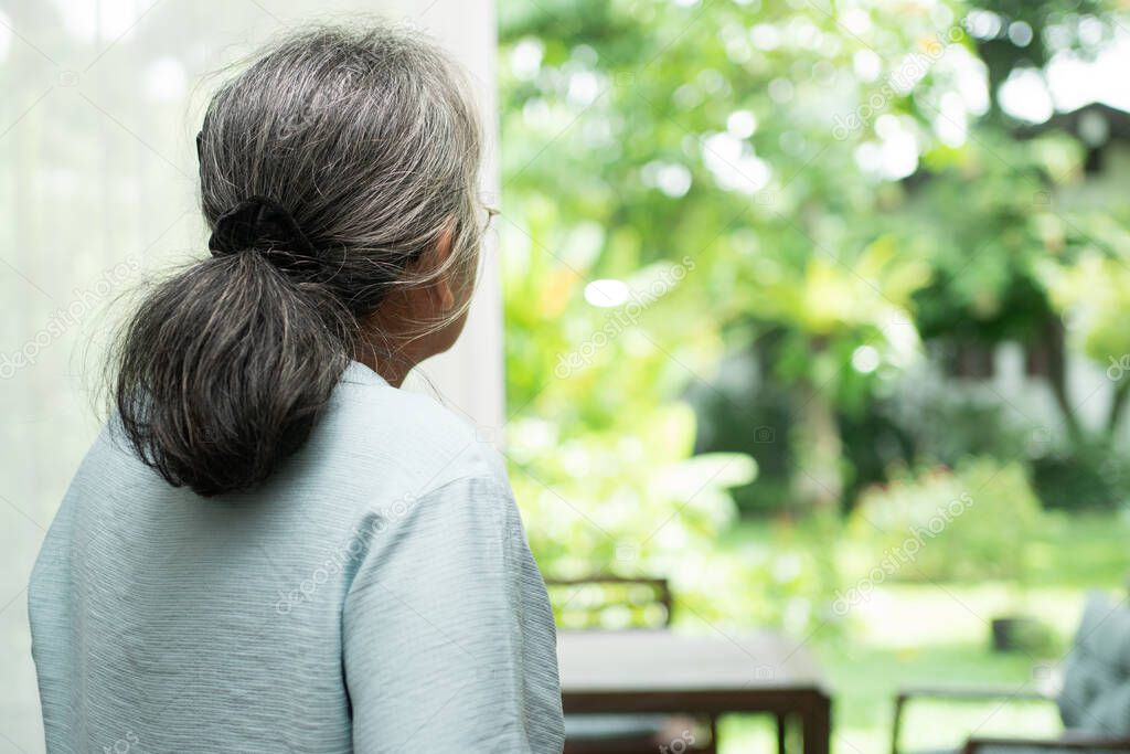 A sad old elderly woman uses Walker for standing in front of windows and looking outside and feeling lonely. Concept of depression caused by illness and Elderly caregivers