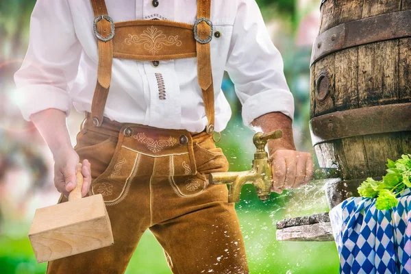 Bavarian man in leather trousers taps a wooden barrel of beer — Stock Photo, Image