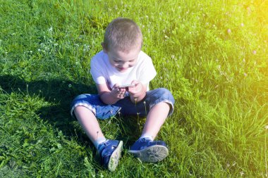 Çocuk yaz aylarında çim üzerinde oturan ve iskambil spor üstünde hareket eden telefon. Arka plan otu ve güneş ışığı