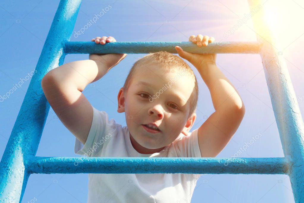 A blond boy holds onto the crossbar on a sports ground in the summer. Close-up portrait of a child