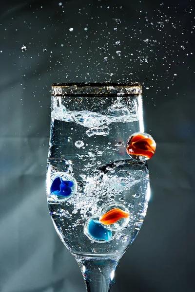 Several glass balls fall into a glass of water and create waves and air bubbles. Splash of water in the glass. One ball fly past the glass.