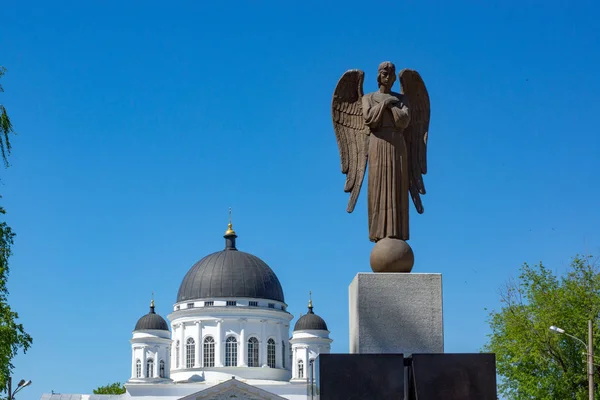 Monument in the form of an angel with folded arms on his chest, wings behind his back. Behind it is a white stone Christian Cathedral with black domes. Against the blue cloudless sky.