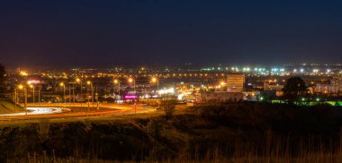 Gece şehir ışıkları. Sokak lambaları ve otomobillerin ışık parçaları tarafından aydınlatılmış yoldan sola dön. Ön planda çim tepeler vardır.