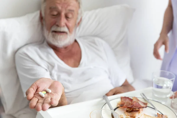 Nurse give medicine pills to elderly senior patient while he is in his bed with breakfast