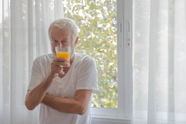 Yaşlı bir emeklinin elinde bir bardak portakal suyu tutarak odanın penceresinin yanında durup mutlulukla dışarı bakması.