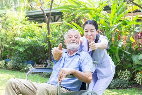 一位护士照顾一位年长的男性 并在自家花园的轮椅上竖起大拇指 — 图库照片