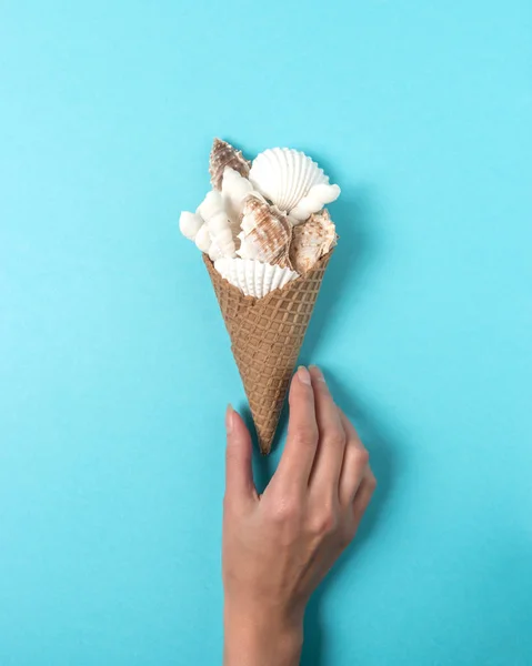 Tvůrčí kompozice s mušlemi a kužel zmrzliny v ruce na pastelově modré pozadí. Letní minimální koncept. — Stock fotografie
