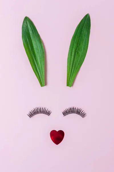 Bunny face made of natural green leaves with eyelashes on pastel pink background. Easter minimal concept. Flat lay.