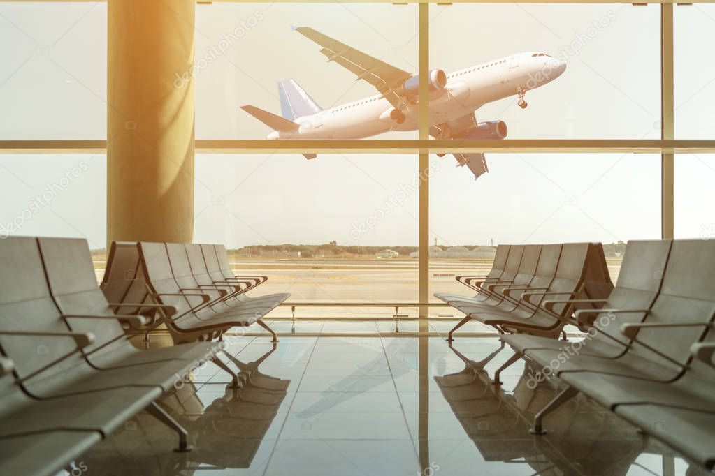 Empty chairs in the departure hall at airport on background of airplane taking off at sunset. Travel concept.