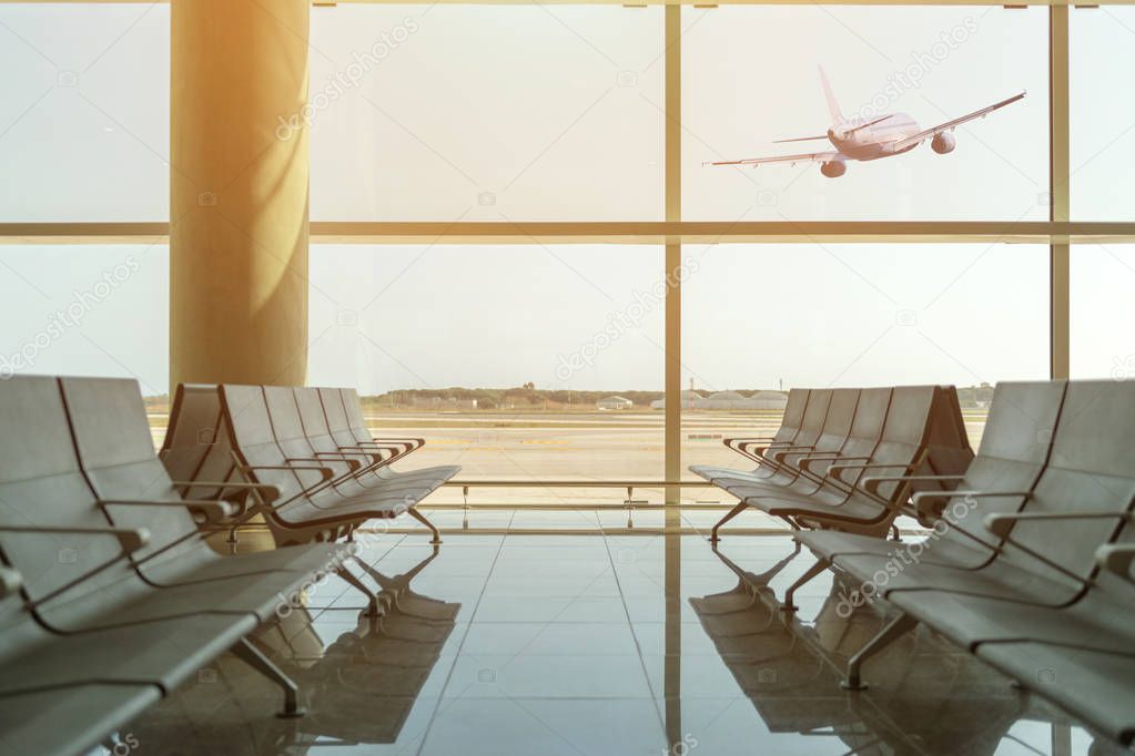 Empty chairs in the departure hall at airport on background of airplane taking off at sunset. Travel concept.