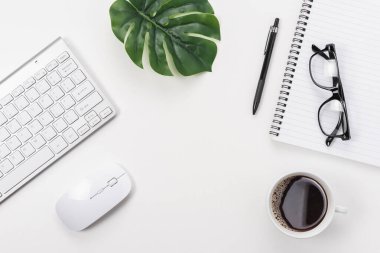 Bilgisayar klavyesi, ofis malzemeleri, yeşil yaprak ve beyaz arka planda kahve fincanı olan çalışma alanı.