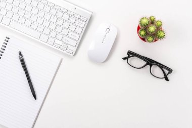 Boş defter, bilgisayar klavyesi ve diğer ofis malzemeleriyle dolu beyaz ofis masası. Kopyalama alanı olan üst görünüm.