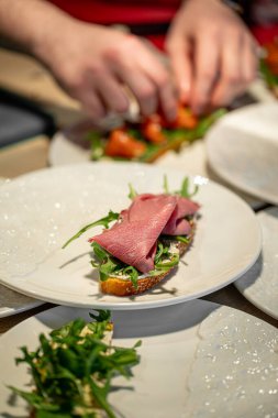 Beyaz tabakta pişmiş jambon ve roka ile bruschetta yapan şefin yakın çekimi.
