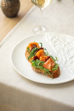 Somonlu gurme bruschetta, avokado soslu ve roka soslu, susamlı baget dilimli, beyaz tabakta servis ediliyor.