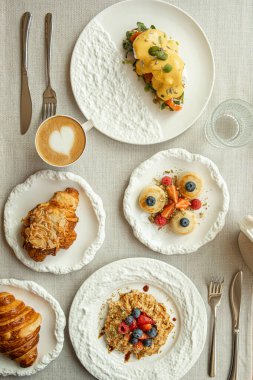 Çeşitli kahvaltı yemekleri, bruschetta, krep ve kapuçinolu kruvasanlar dahil zarif beyaz tabaklar.