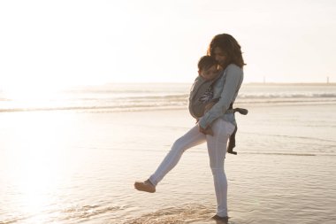 Sahilde yürümeye başlayan çocuk taşıyan ergobaby annesiyle