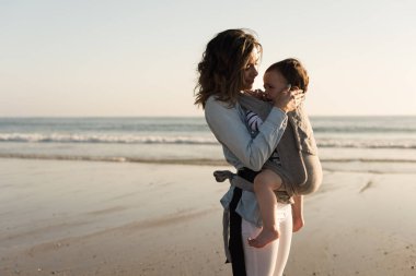 Sahilde yürümeye başlayan çocuk taşıyan ergobaby annesiyle
